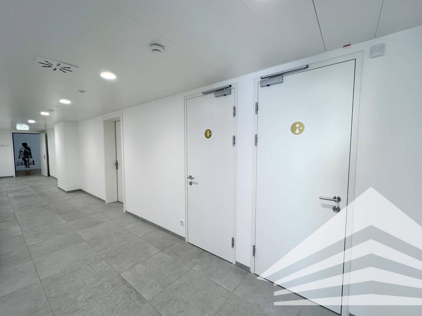 Heller Flur mit Fliesenboden und mehreren Türen zu Toilettenbereichen in einem modernen Gebäude.
