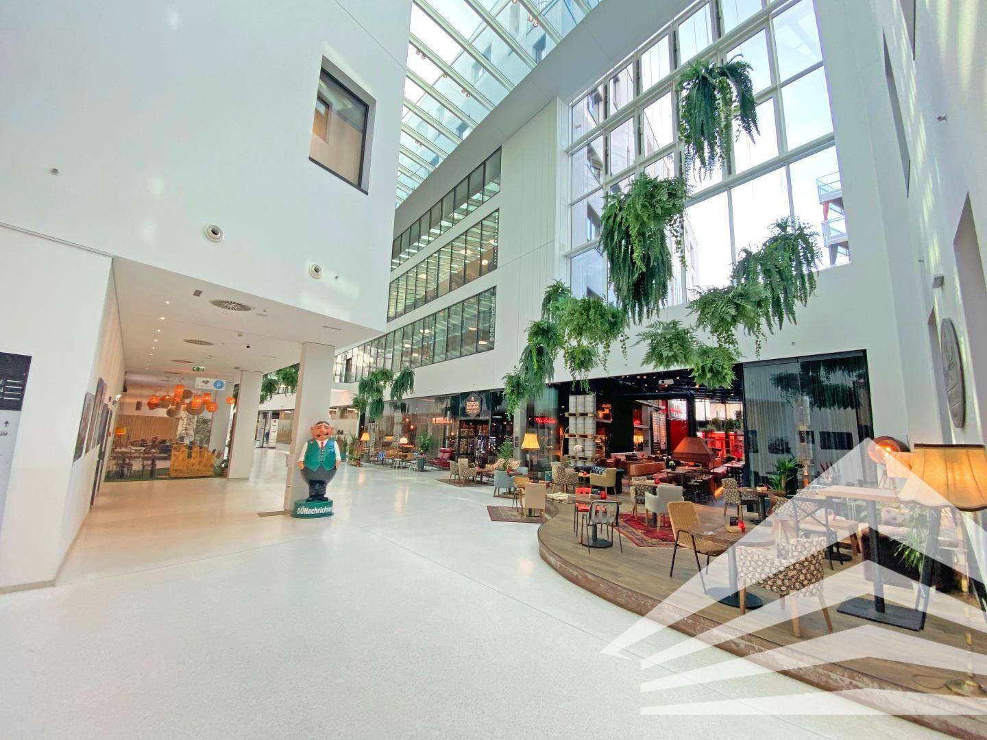 Großzügige, lichtdurchflutete Lobby mit hohen Decken, Geschäften und hängenden Pflanzen in einem modernen Gebäude.