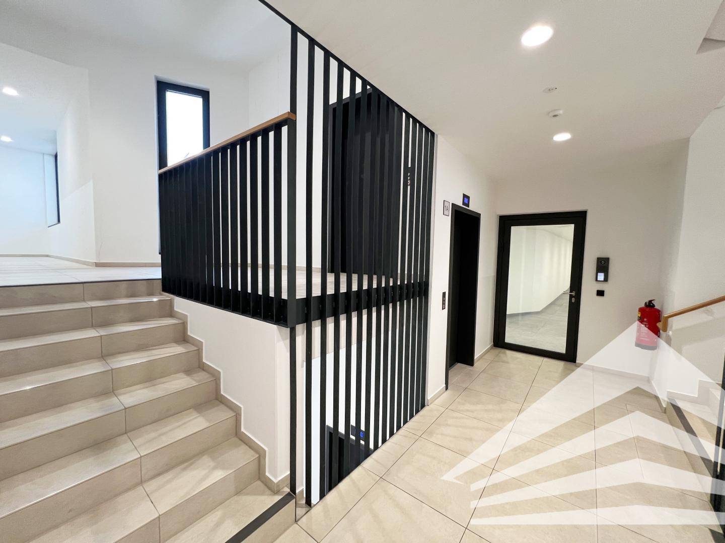 Modernes Treppenhaus mit schwarzem Geländer, Fliesenboden und Aufzugzugang in einem Geschäftsgebäude.