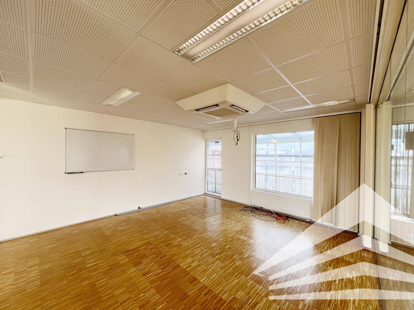 Heller Büroraum mit Parkettboden, einer großen Fensterfront und einer Whiteboard-Wand für Präsentationen.