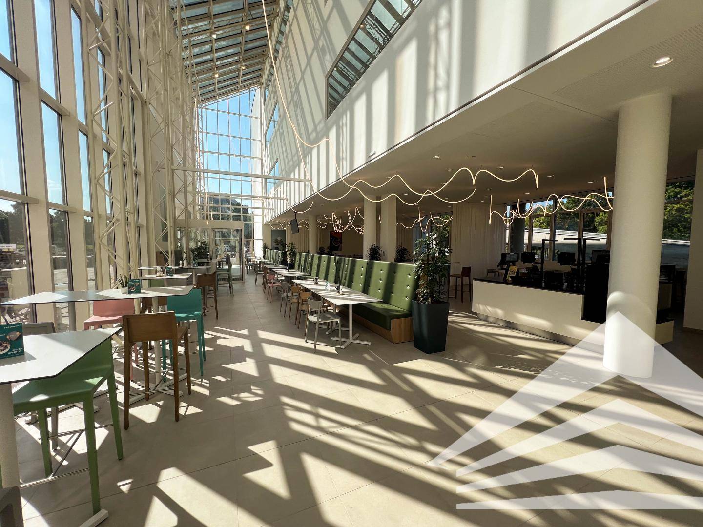 Heller, moderner Cafeteria-Bereich mit hohen Glaswänden, farbigen Stühlen und viel natürlichem Licht.