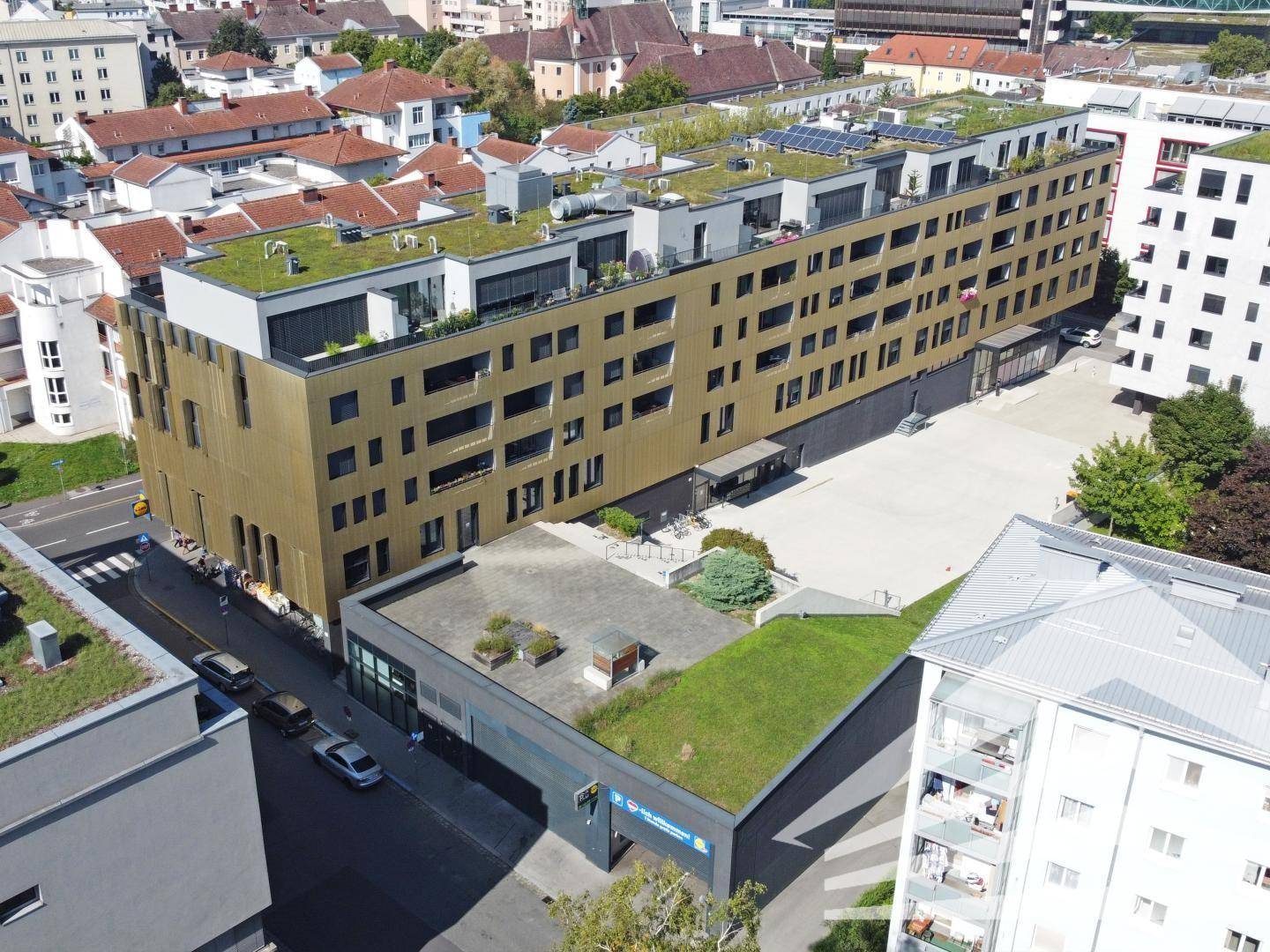 Luftaufnahme eines modernen Gebäudekomplexes mit Gründächern und Photovoltaikanlagen in städtischer Umgebung.