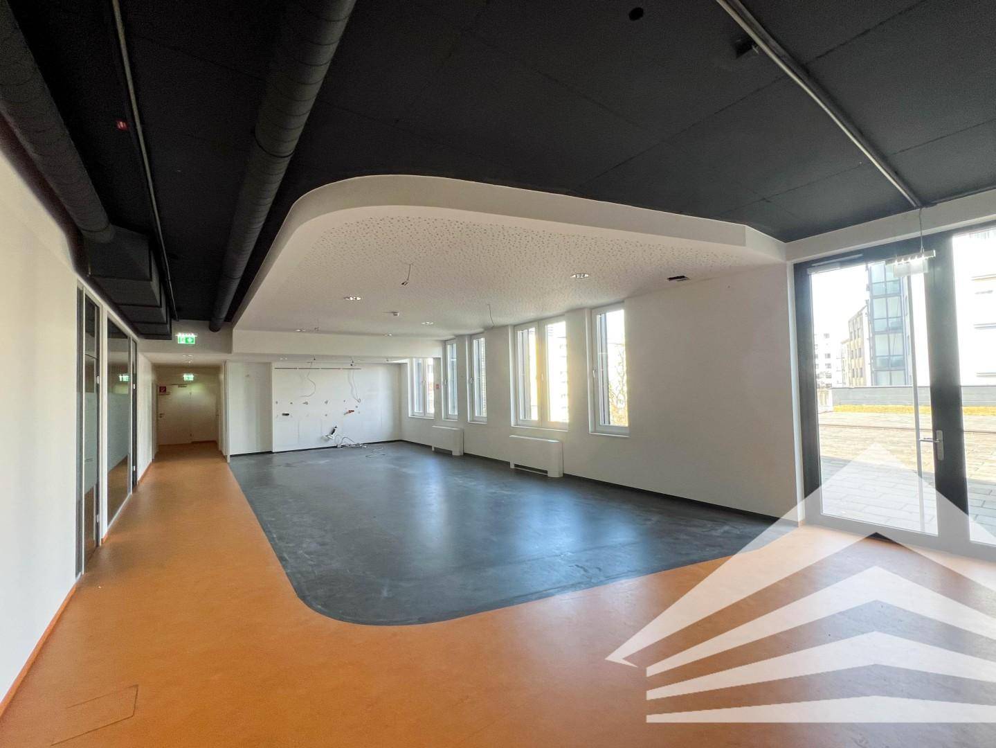 Großer, leerer Büroraum mit vielen Fenstern, dunklem Bodenbelag und moderner Deckenkonstruktion.