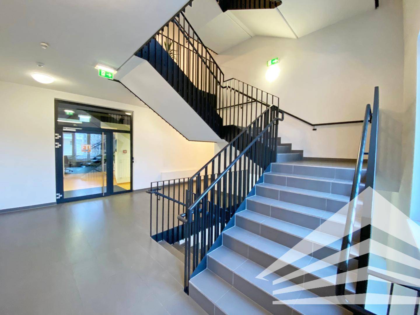 Modernes Treppenhaus mit grauen Stufen, schwarzen Geländern und einem hellen Eingangsbereich.