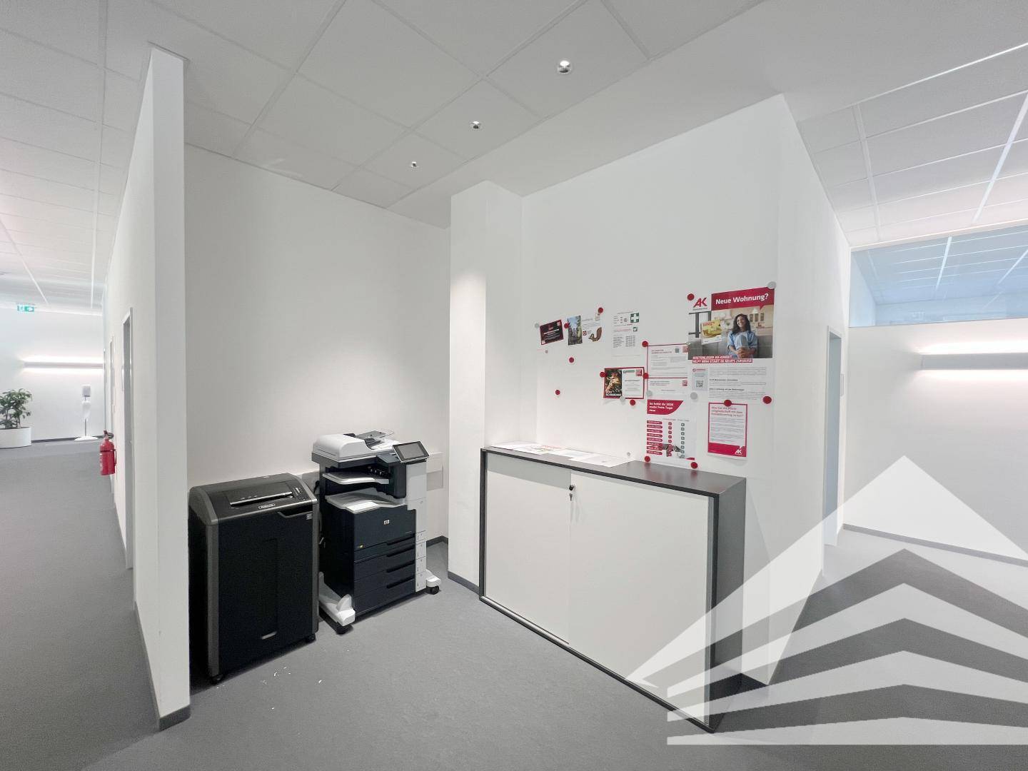 Heller Bürogang mit Druckerstation, Ablageschränken und Informationsmaterial an der Wand.