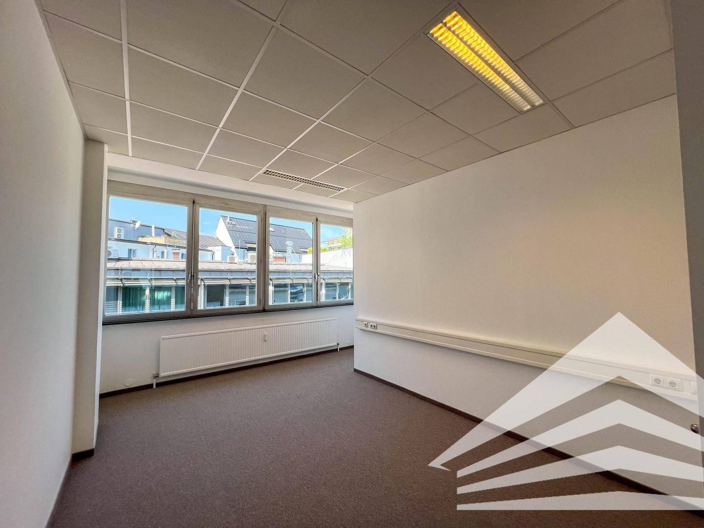 Büro mit Teppichboden, Fensterfront, Kabelkanälen an der Wand und viel natürlichem Licht.