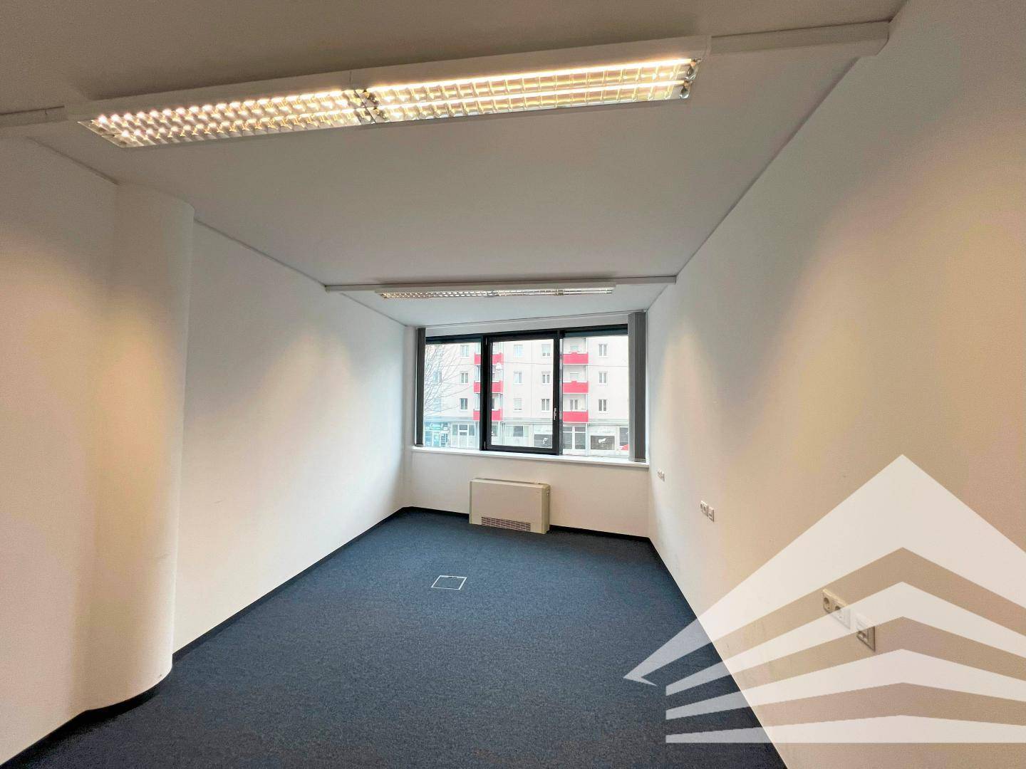 Kleinerer, leerer Büroraum mit Teppichboden, einem Fenster und funktionaler Deckenbeleuchtung, bereit zur Nutzung.