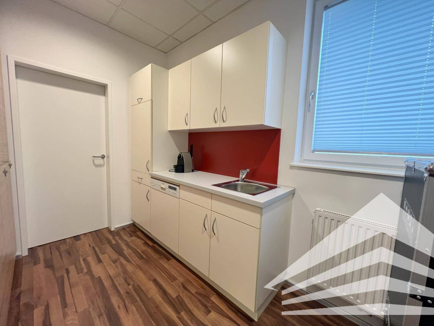 Funktionale Teeküche mit weißen Schränken, Spülbecken und Fenster mit Jalousien.