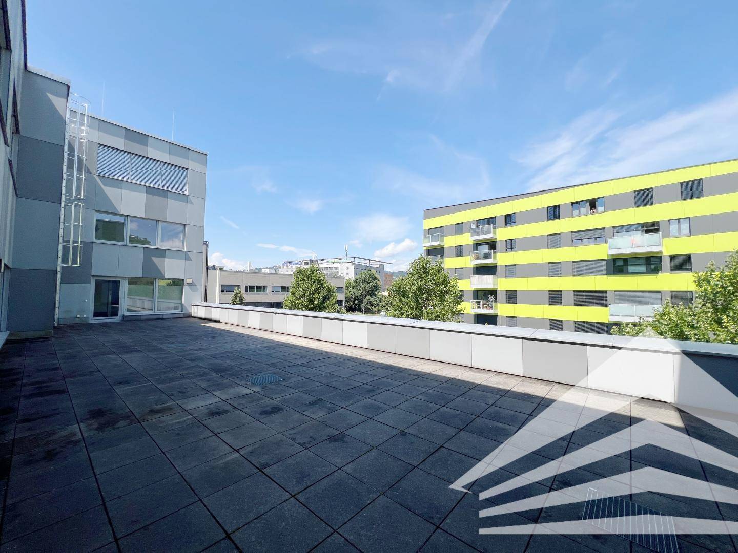 Große Außenterrasse mit grauen Pflastersteinen, umgeben von modernen Bürogebäuden.