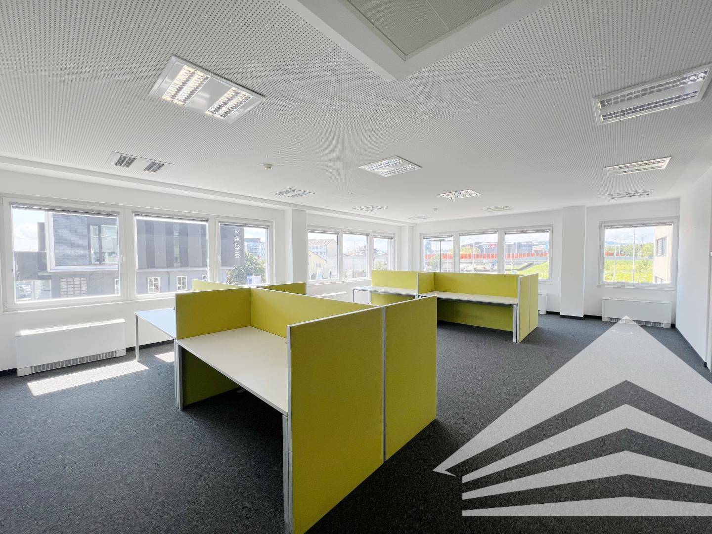 Großraumbüro mit mehreren Arbeitsplätzen, gelben Trennwänden und vielen Fenstern für Helligkeit.