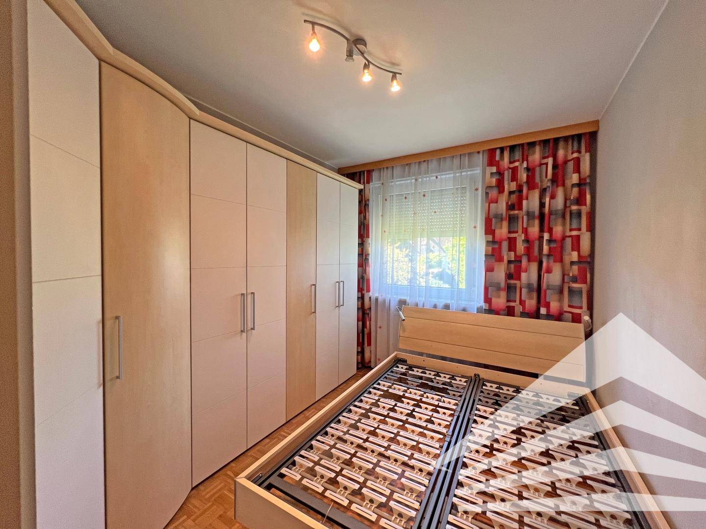 Schlafzimmer mit Bettgestell, großem Einbauschrank und Fenster mit roten Vorhängen.