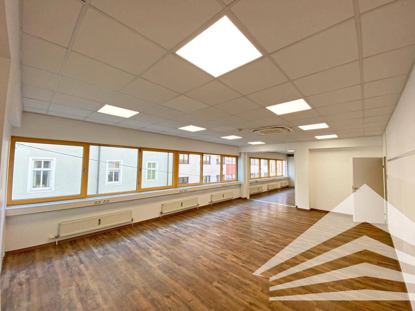 Helles Büro mit langen Fensterfronten, Laminatboden und Deckenbeleuchtung.