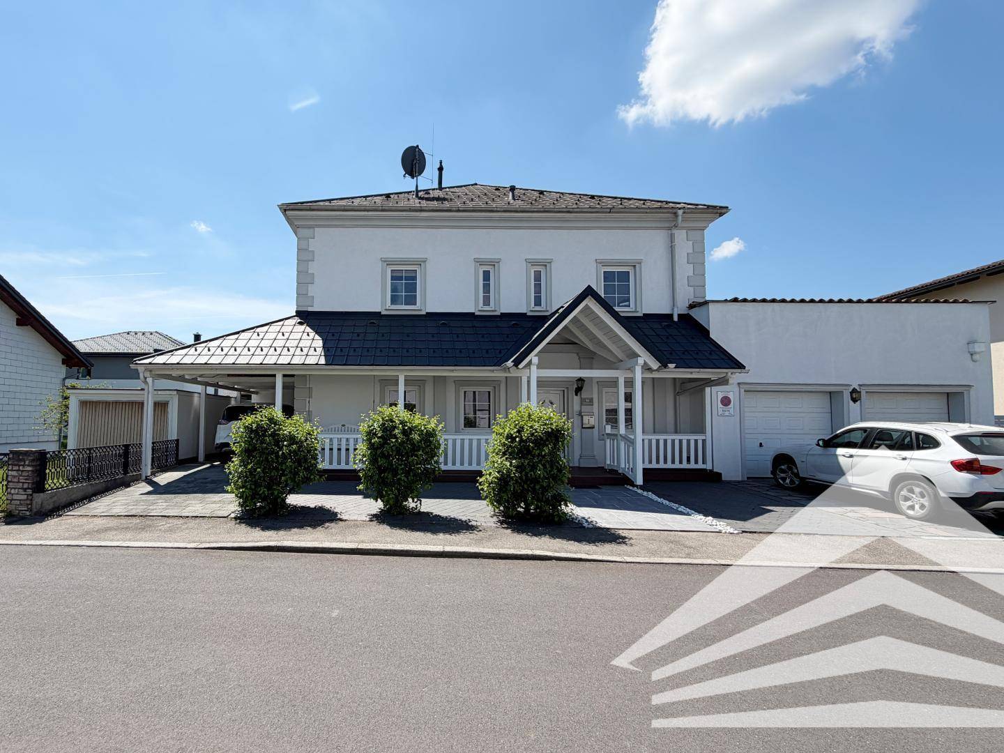 Vorderansicht des weißen Hauses mit Veranda, Einfahrt und Carport unter klarem Himmel.
