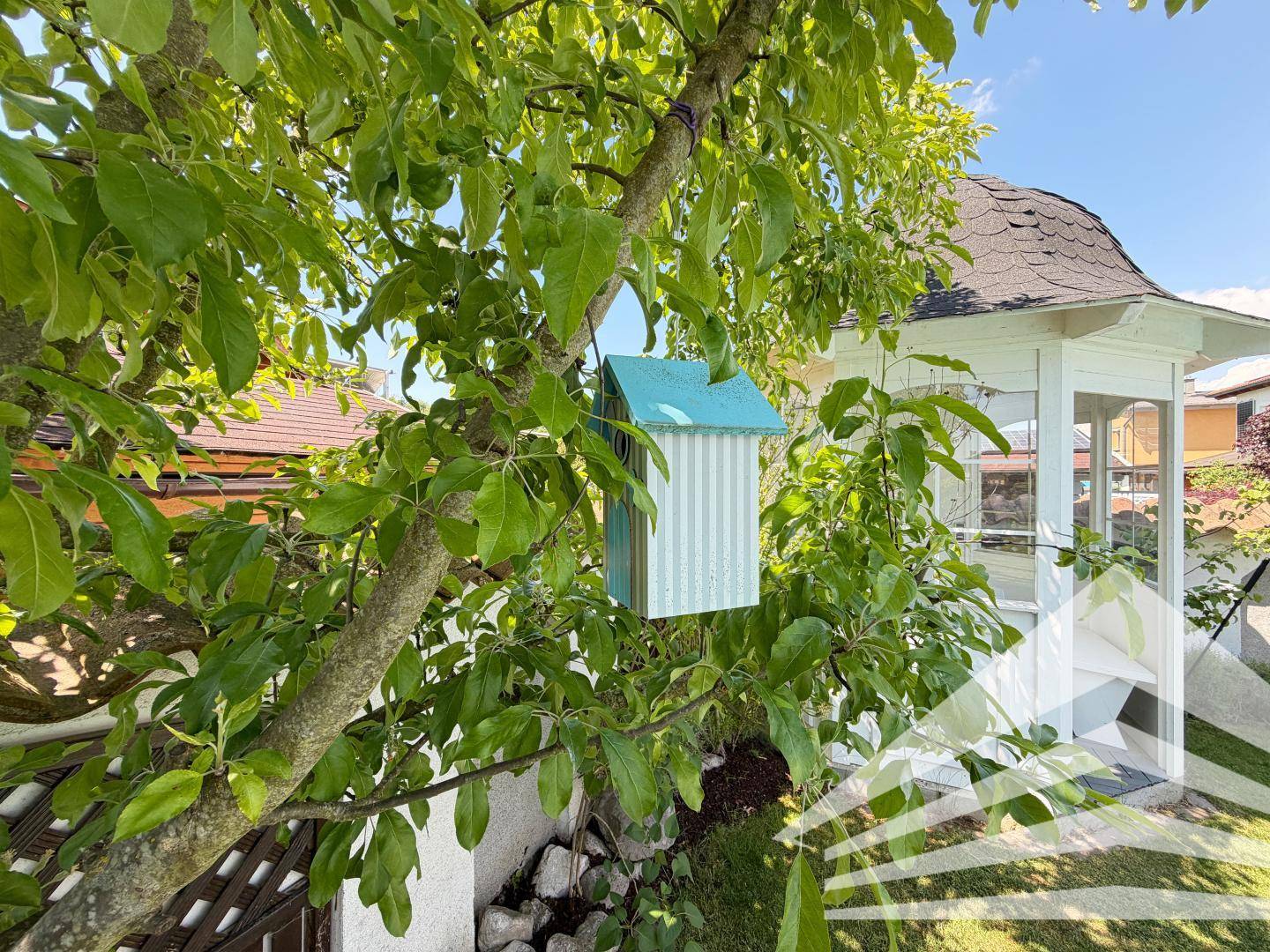 Charmantes Vogelhaus hängt an einem Baum, mit einem weißen Gartenpavillon im Hintergrund.