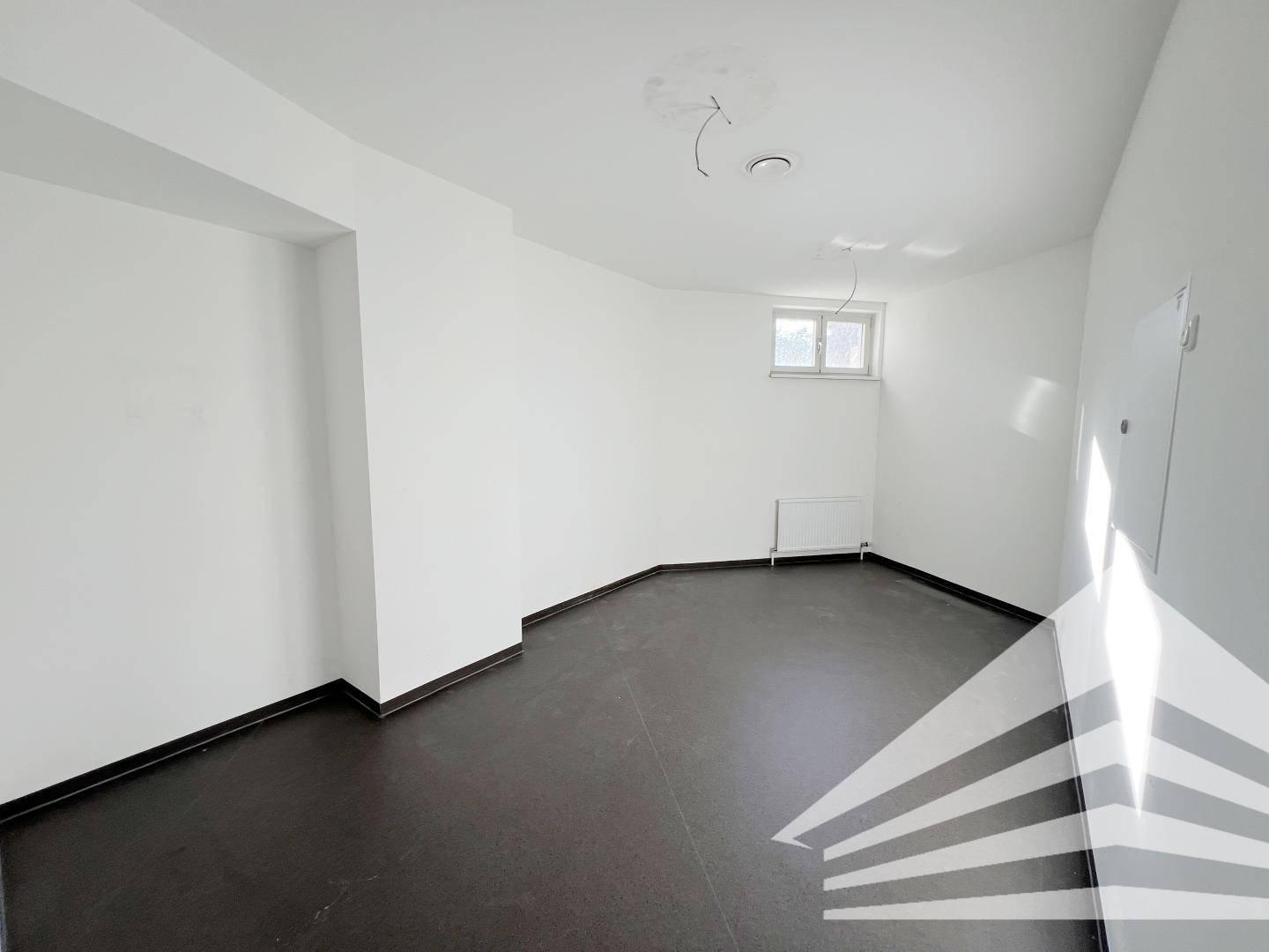 Kleiner, leerer Raum mit weiß gestrichenen Wänden, dunklem Boden, einem Fenster und einem Heizkörper.
