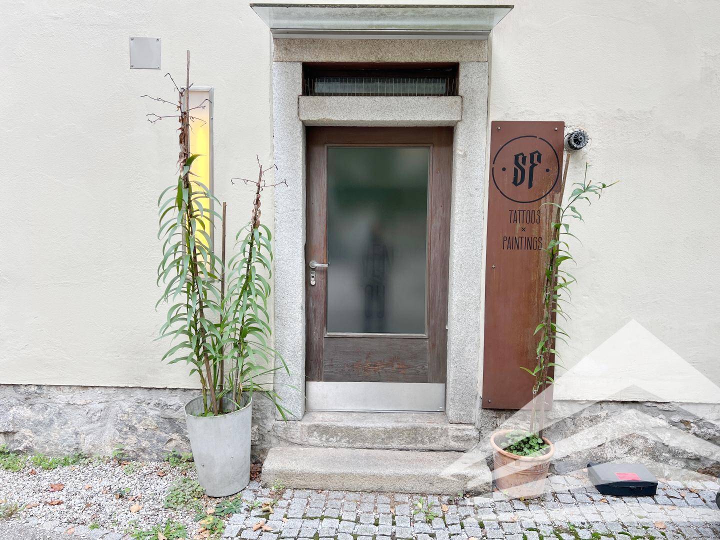 Eingangsbereich eines Geschäfts mit Holztür, Steinrahmen und Pflanzendekoration.