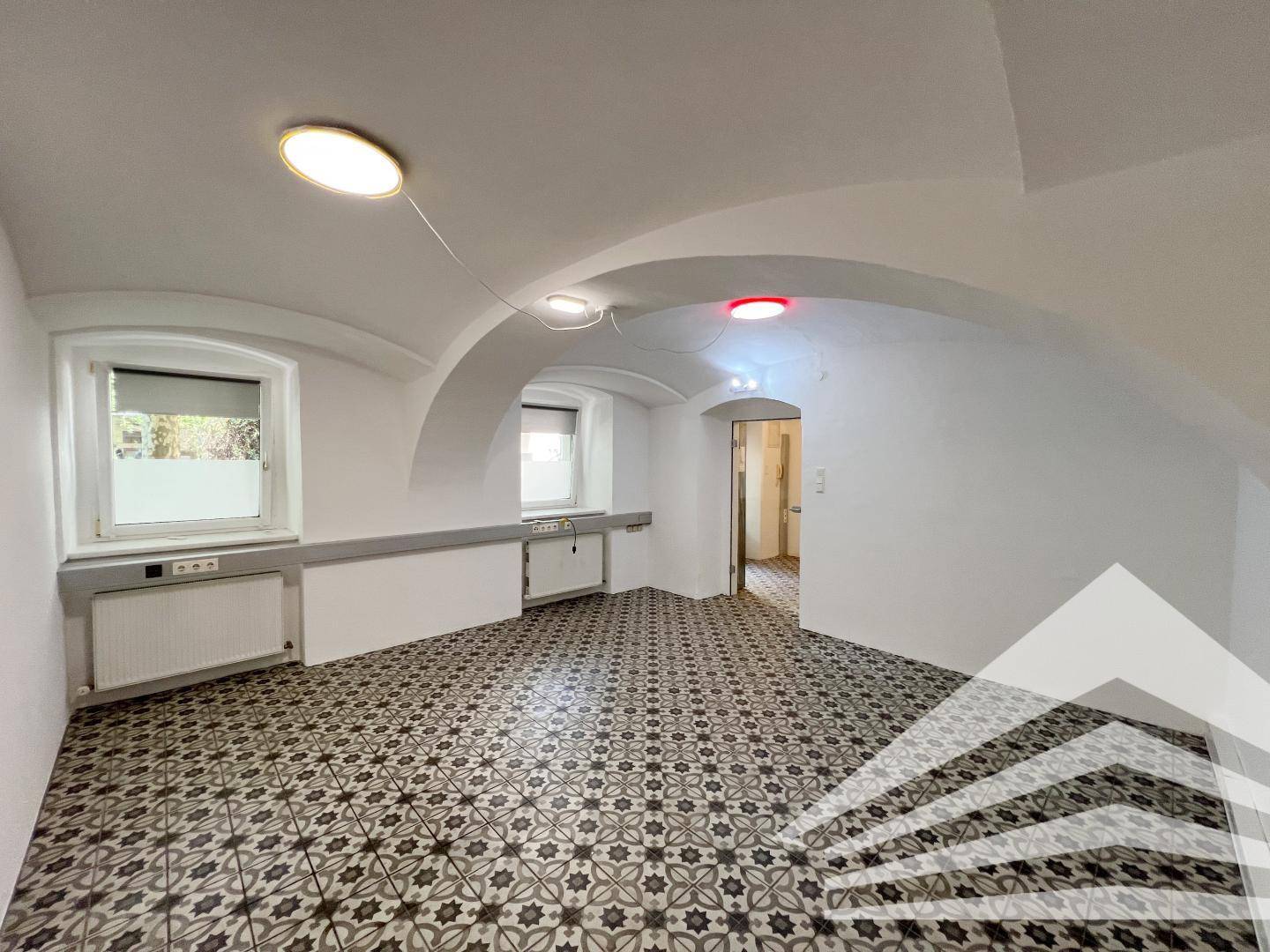 Großer Raum mit zwei Fenstern, Gewölbedecke und auffälligem Fliesenboden.