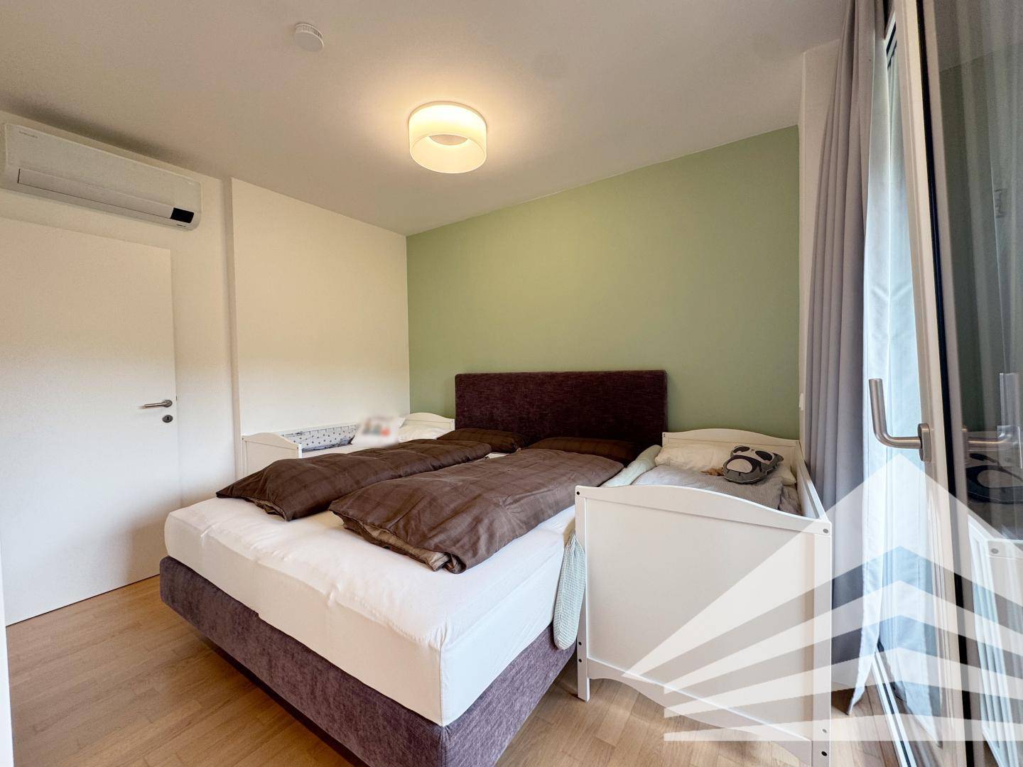 Schlafzimmer mit Doppelbett, grüner Akzentwand, Holzboden und Klimaanlage.
