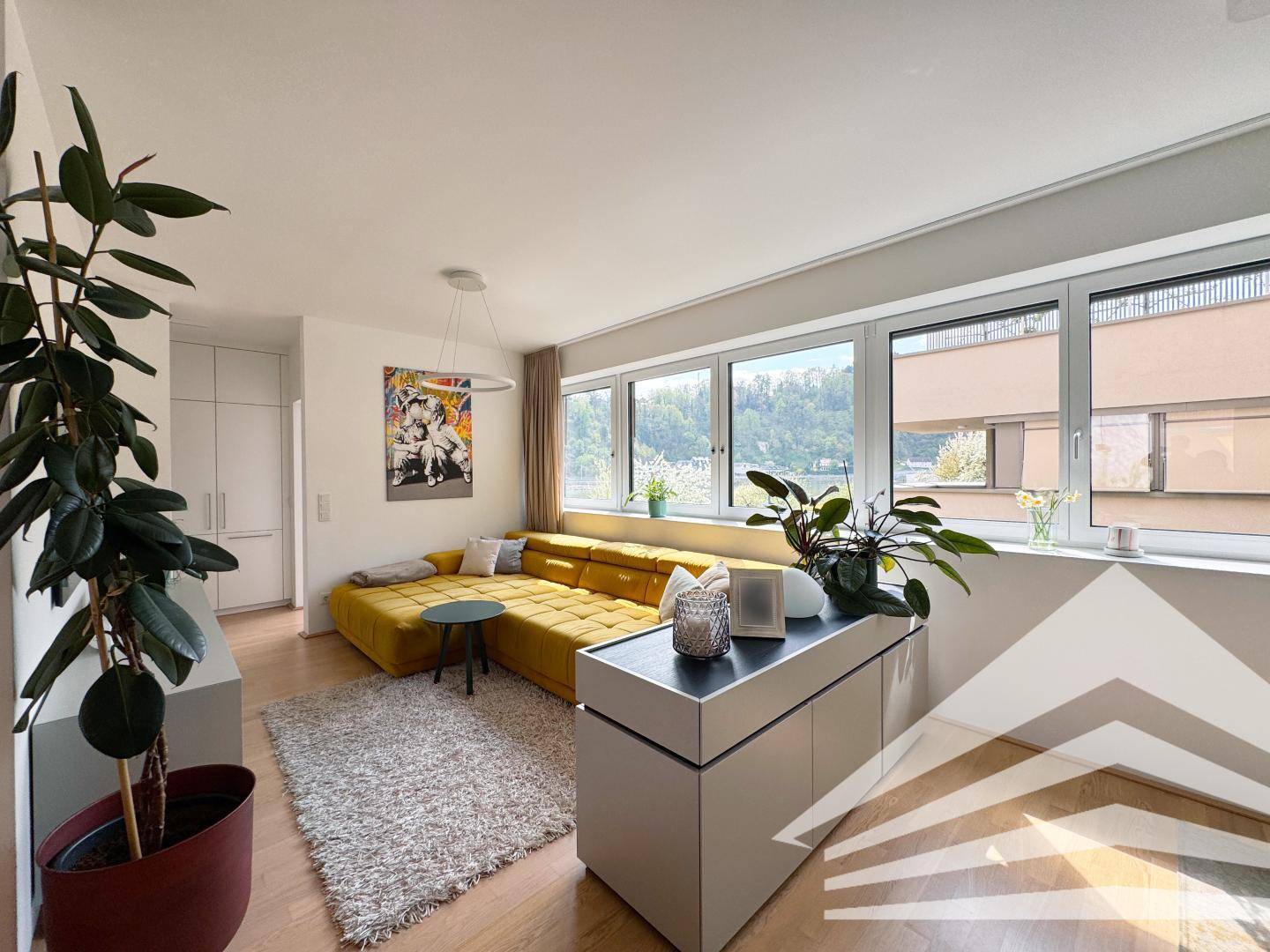 Helles Wohnzimmer mit gelbem Ecksofa, Holzboden und großen Fenstern mit Blick ins Grüne.