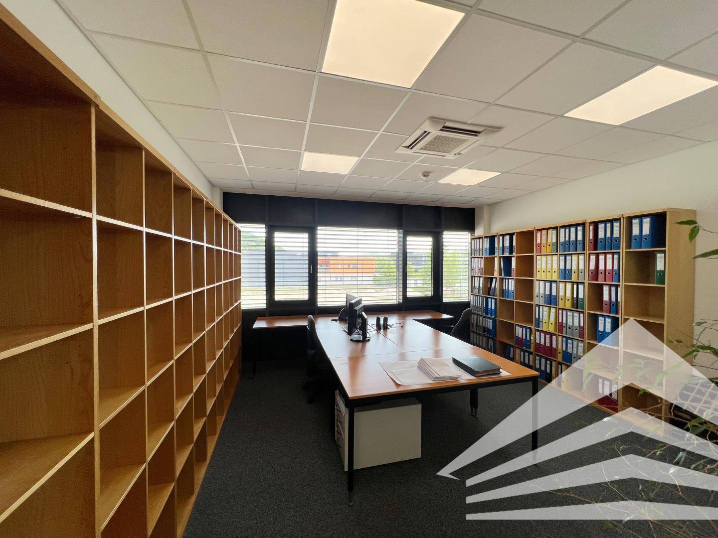 Geräumiges Büro mit großen Regalen, Schreibtisch und Fenstern mit Jalousien für viel Tageslicht.