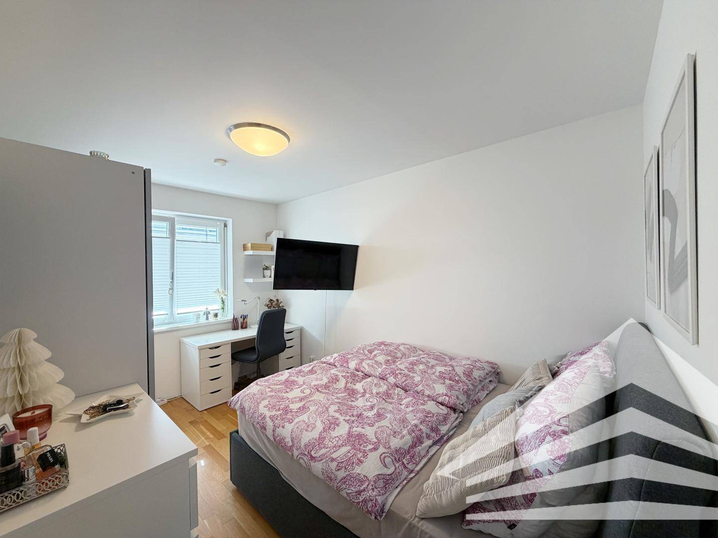 Gemütliches Schlafzimmer mit Holzboden, Schreibtisch am Fenster und Wandfernseher.