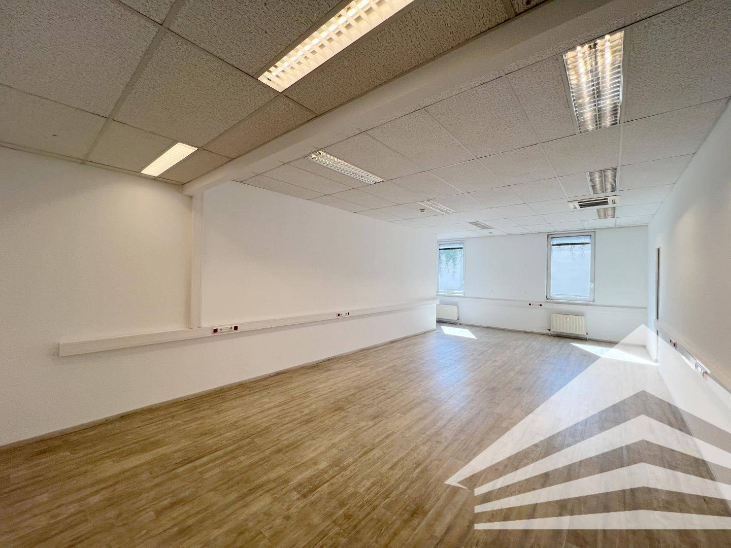 Leeres Büro mit weiß gestrichenen Wänden, Laminatboden und zwei Fenstern, die für ausreichend Helligkeit sorgen.