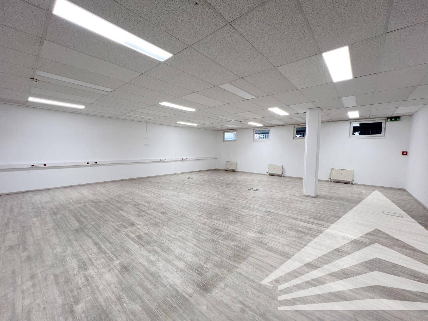 Großes, leeres Büro mit modernem grauem Laminatboden und mehreren kleinen Fenstern, die den Raum erhellen.
