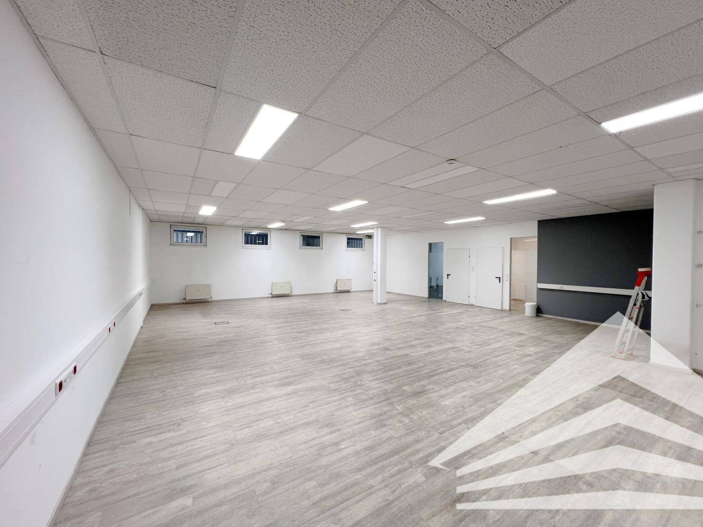 Weitläufiges Büro mit grauem Laminatboden, weißen Wänden und einer dunklen Akzentwand im Hintergrund.