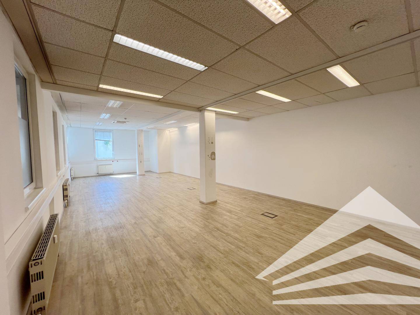 Geräumiges Büro mit einem tragenden Pfeiler, hellem Laminatboden und Fenstern, die den Raum mit Licht füllen.