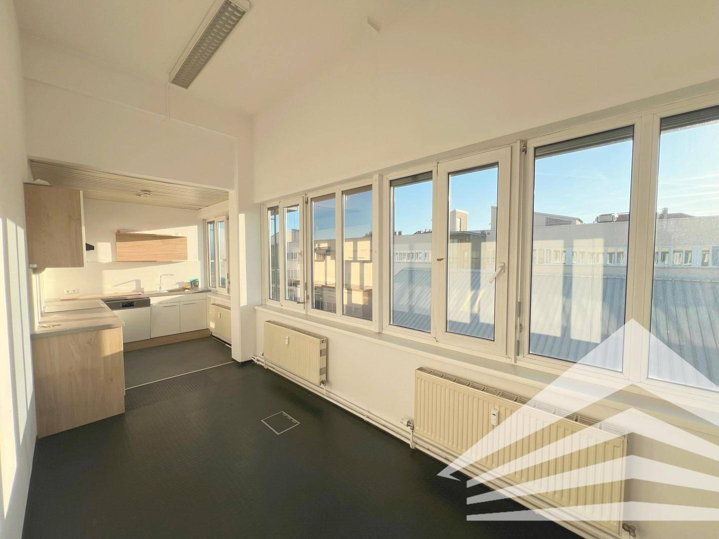 Büroraum mit integrierter Teeküche, dunklem Boden und breiter Fensterfront für natürliches Licht.