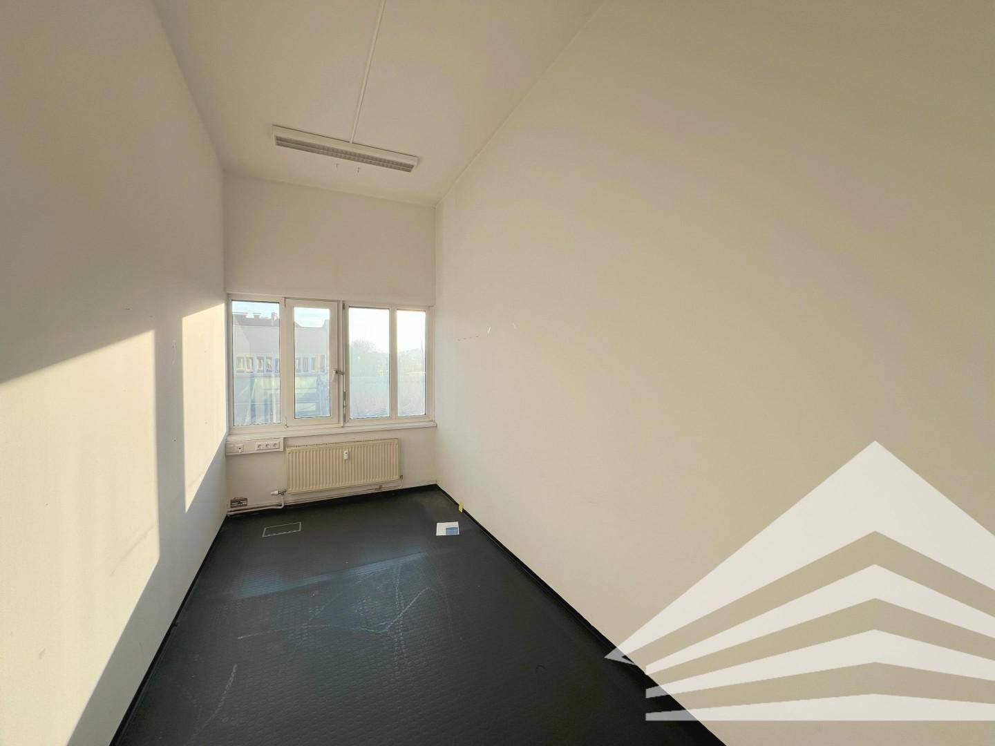 Kleinerer Büroraum mit weißen Wänden, dunklem Boden und drei Fenstern, die Sonnenlicht hereinlassen.