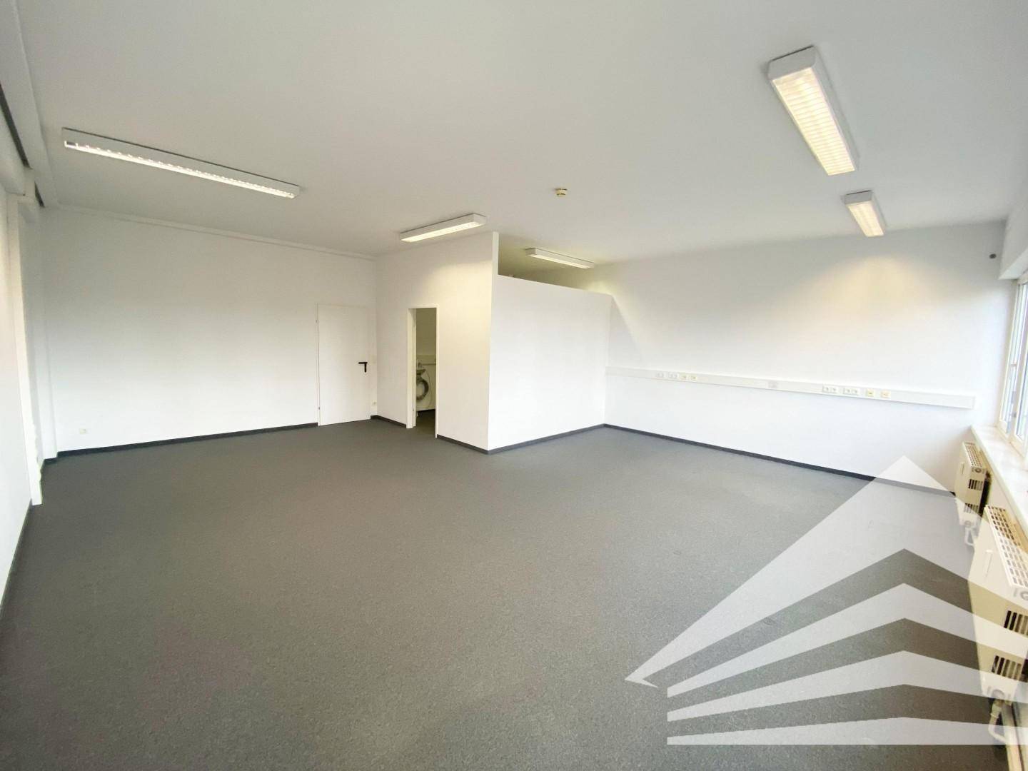 Heller Büroraum mit Teppichboden, weißen Wänden und Zugang zu einem kleinen Nebenraum mit Toilette.