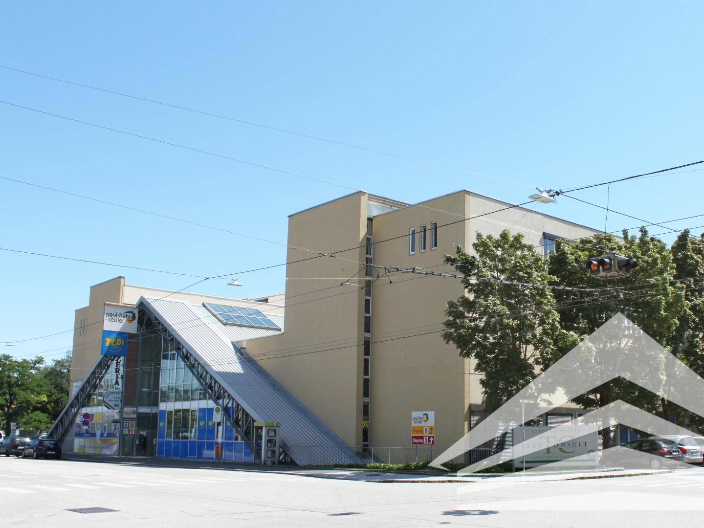 Modernes Geschäftsgebäude an einer Straßenecke mit markanter Glasfassade und Photovoltaikanlage auf dem Dach.