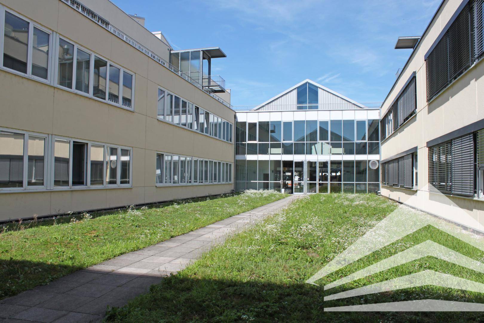Innenhof eines modernen Bürogebäudes mit Glasfassade und einem begrünten Weg, unter blauem Himmel.