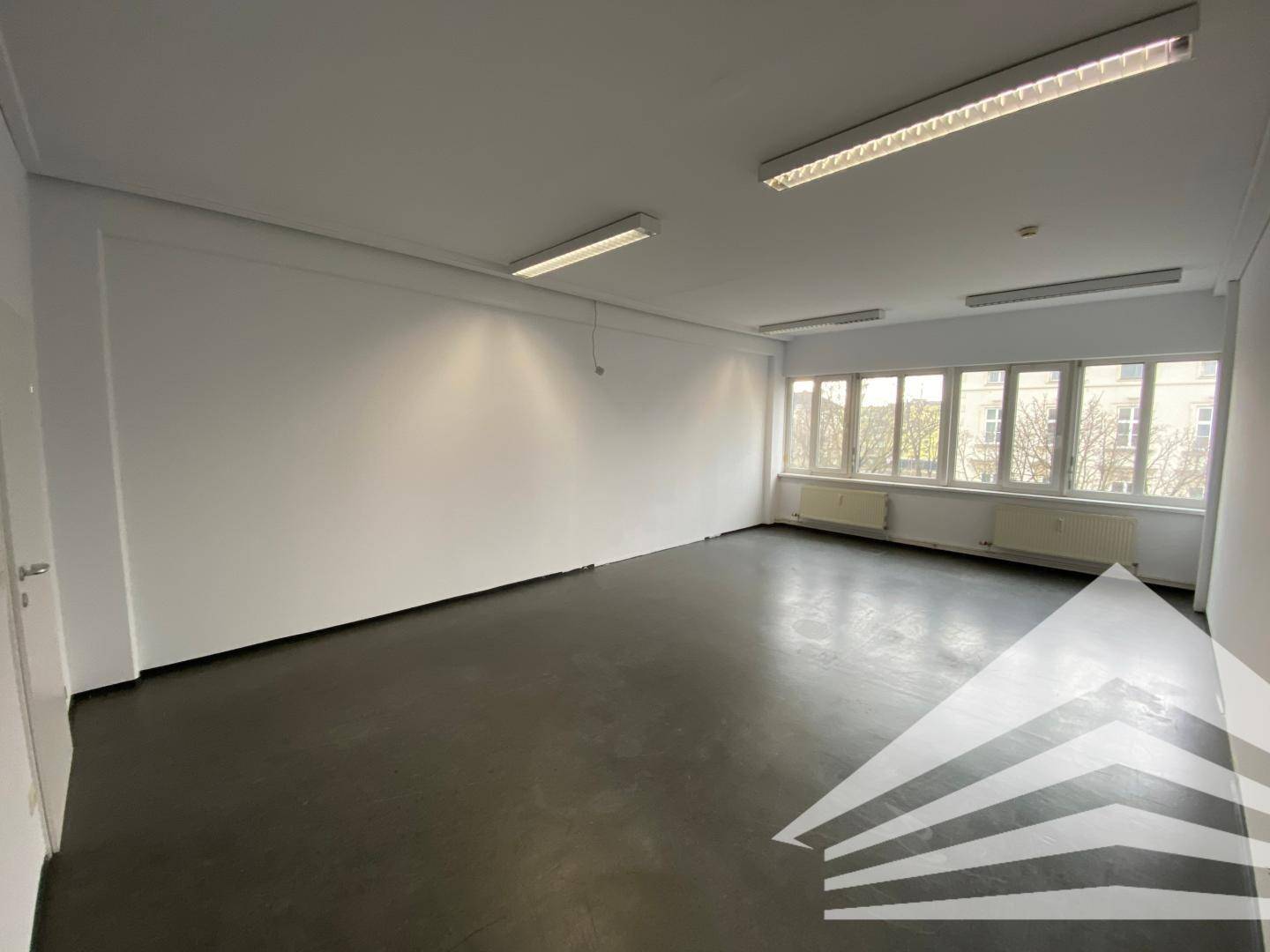 Heller Büroraum mit großen Fenstern, die viel Tageslicht hereinlassen, und einem dunklen Bodenbelag.