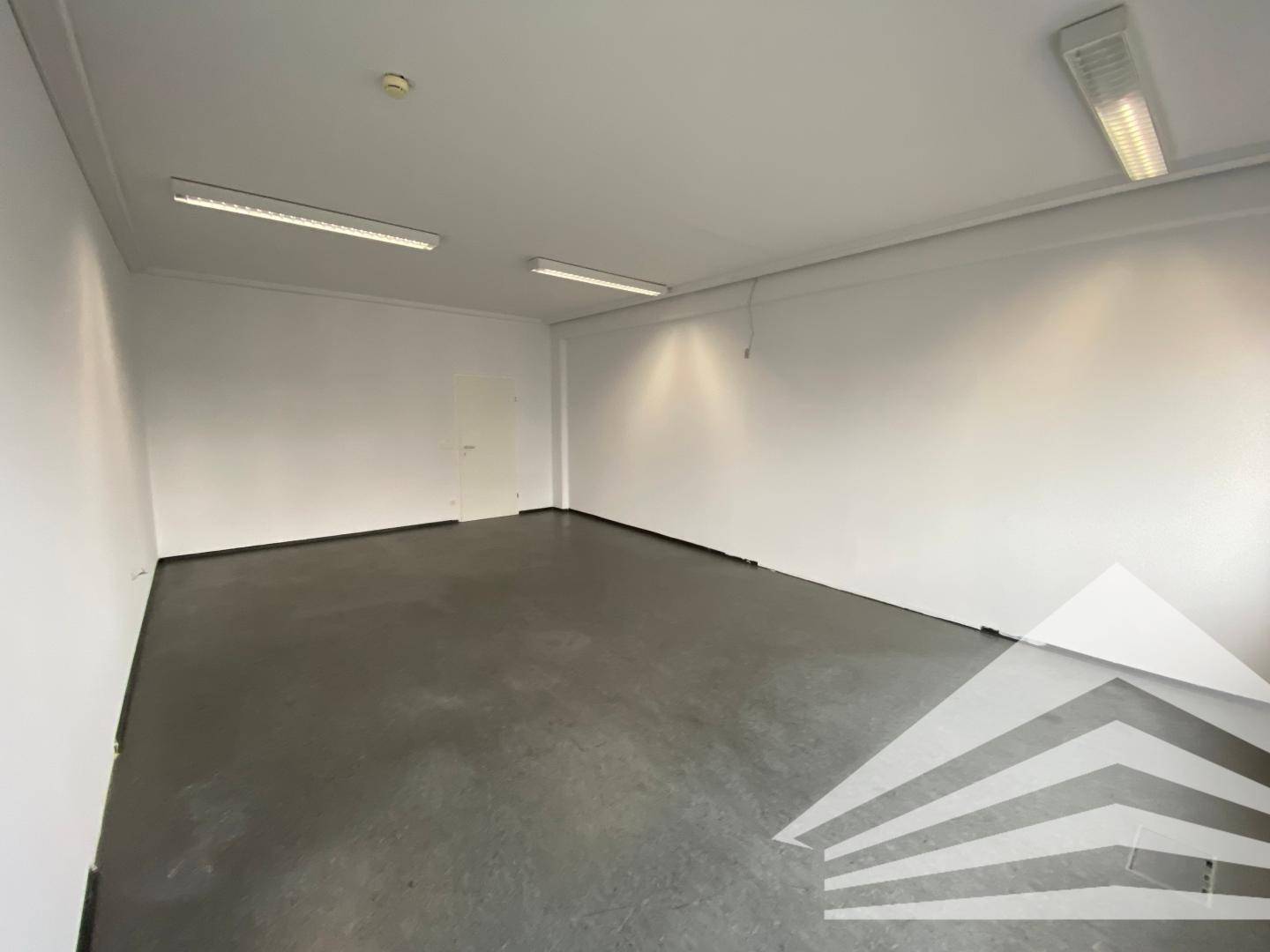 Geräumiger, leerer Büroraum mit weißen Wänden und dunklem Boden, ausgestattet mit Deckenbeleuchtung.