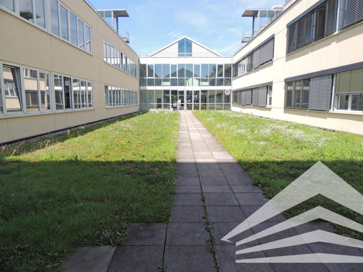 Innenhof zwischen Gebäudeteilen mit einem gepflasterten Weg, der zu einem gläsernen Eingang führt.