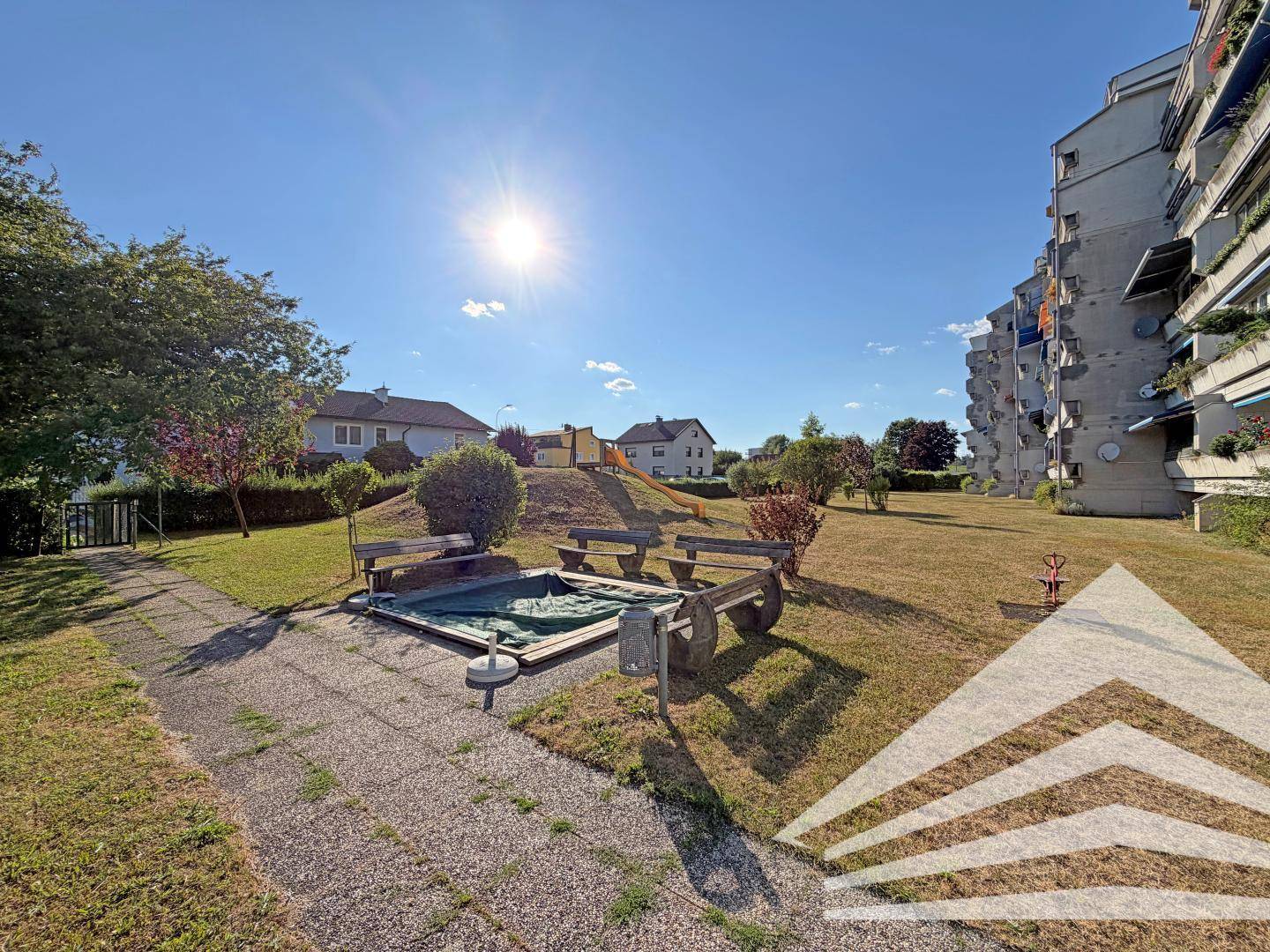 Sonniger Gemeinschaftsgarten mit Sandkasten und Rutsche, umgeben von Rasenflächen und Bäumen.