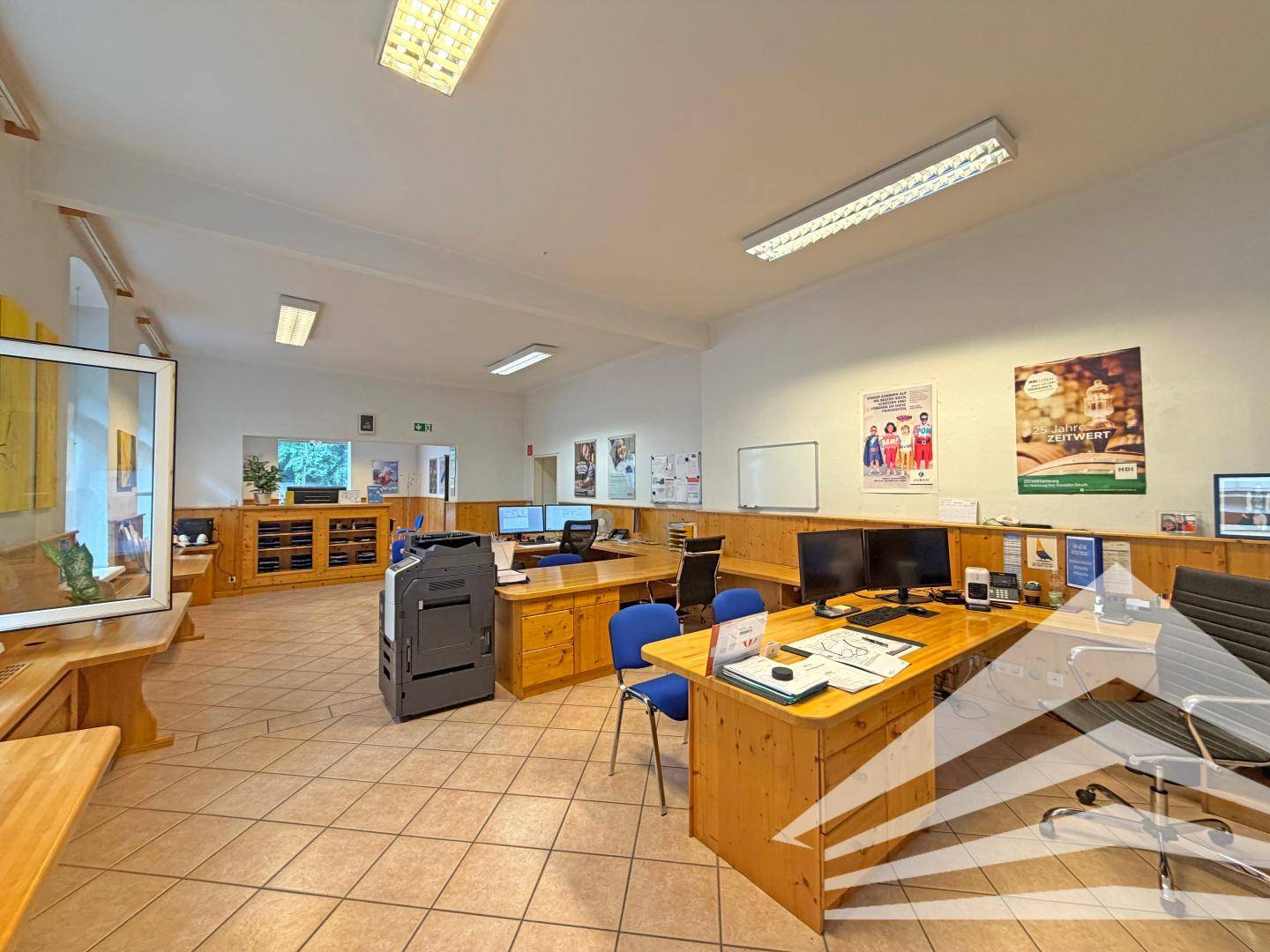 Weitläufiges Büro mit mehreren Schreibtischen, Holzmöbeln und Fliesenboden, ideal für Teamarbeit.