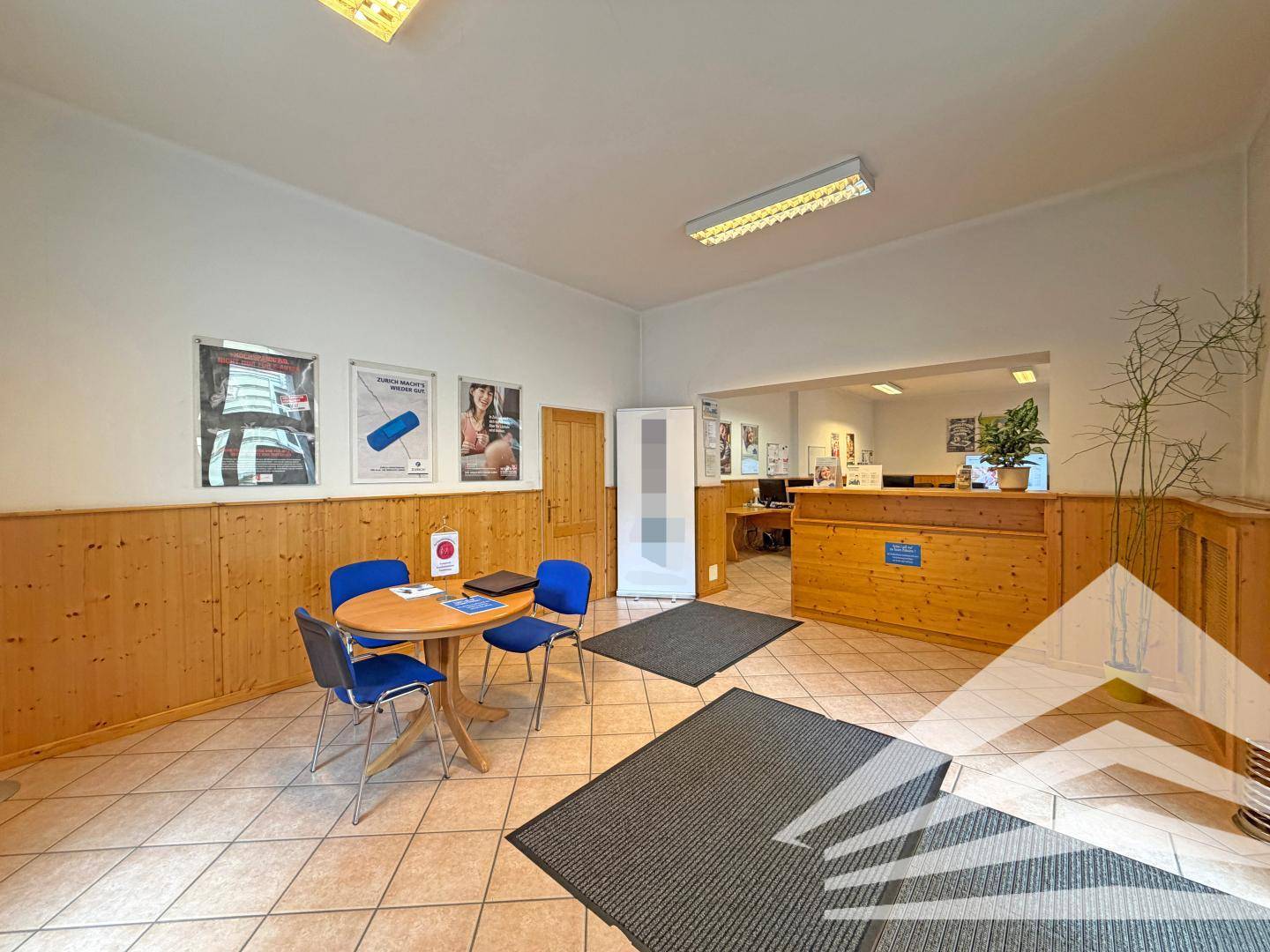 Freundlicher Empfangsbereich mit kleinem Tisch und Stühlen, Holzvertäfelung und Blick in den Bürobereich.