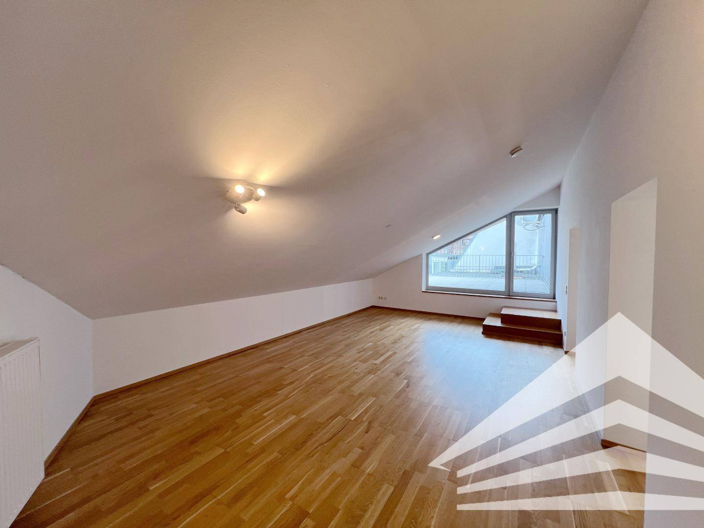 Geräumiger Raum mit Holzboden, Dachschrägen und einem großen Fenster, das viel Licht hereinlässt.