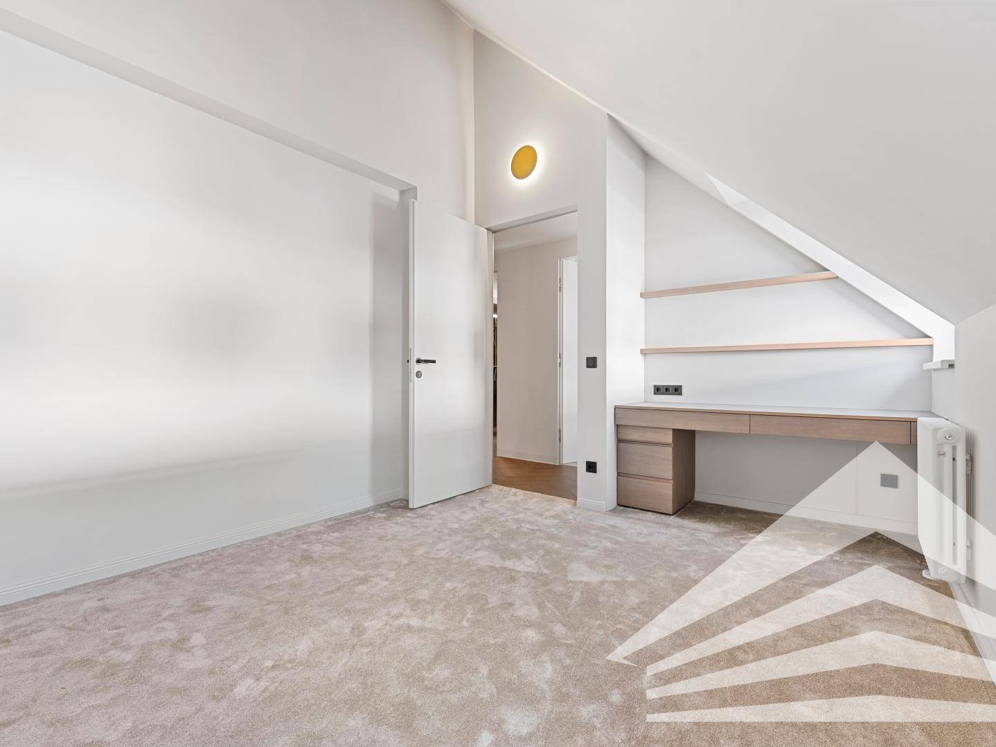 Helles Zimmer mit Dachschräge, Teppichboden und integriertem Schreibtischbereich.