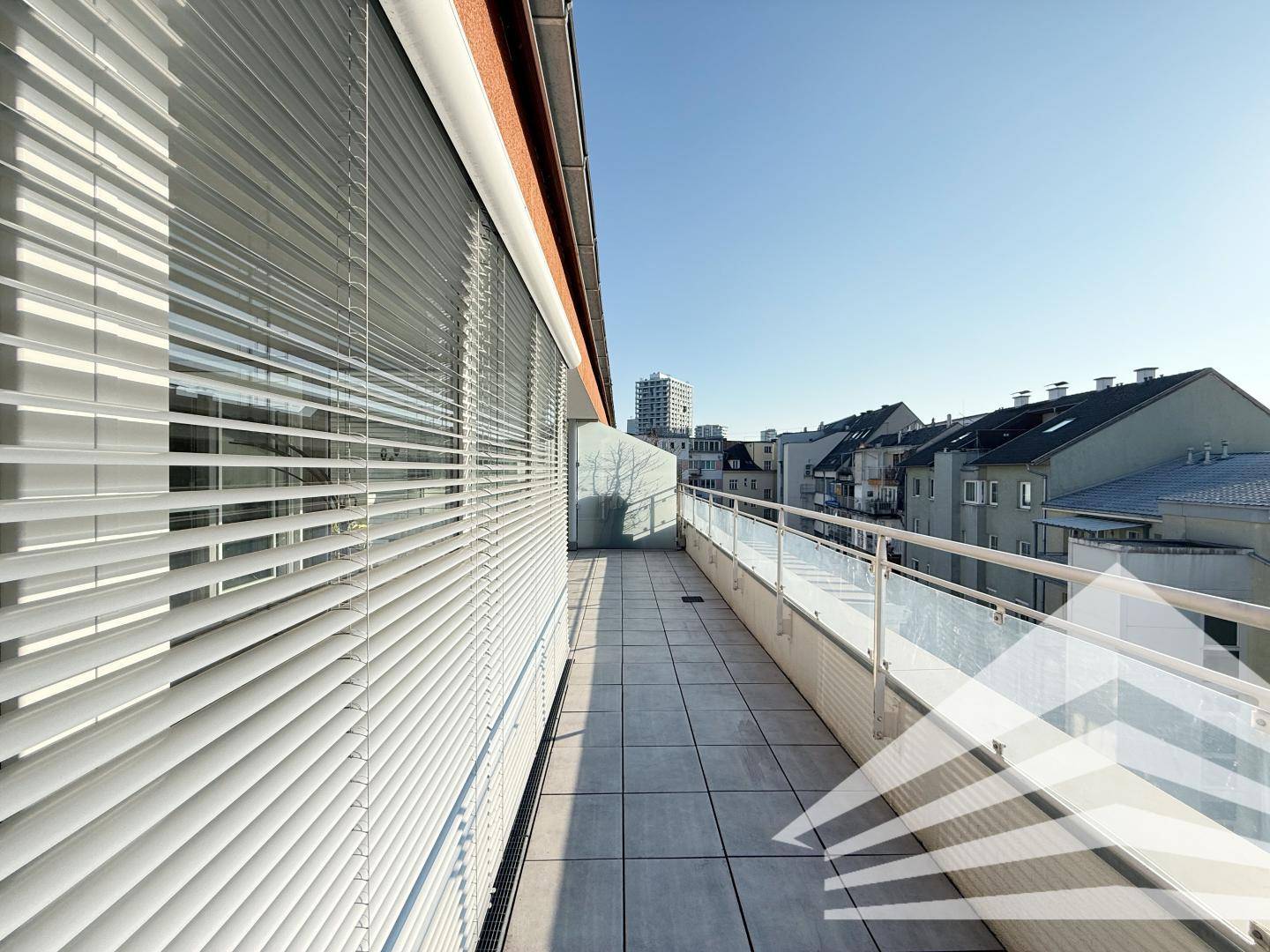 Langer Balkon mit Fliesenboden und Jalousien, bietet einen weiten Blick über die Dächer der Stadt.