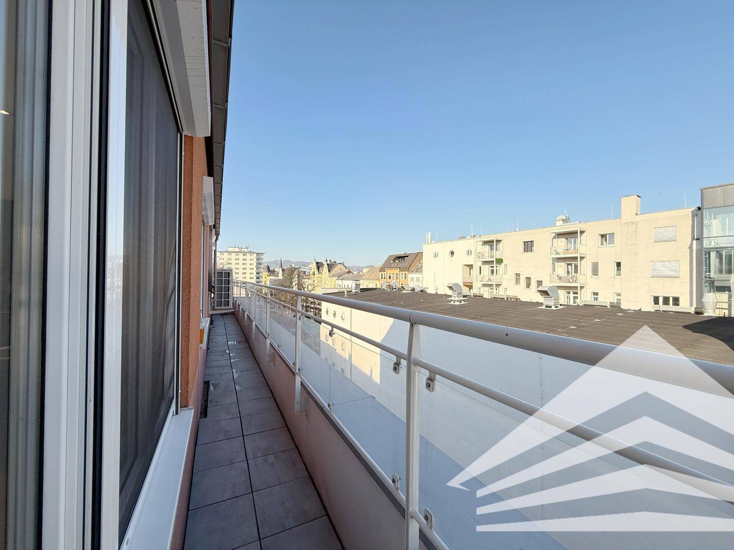Langer Balkon mit Fliesenboden und Glasgeländer, bietet einen weiten Blick über die Stadt.