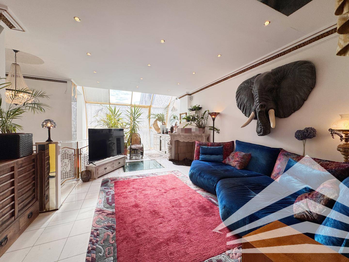 Geräumiges Wohnzimmer mit blauer Samtcouch, großem Teppich und dekorativer Elefantenkopf-Wandskulptur.