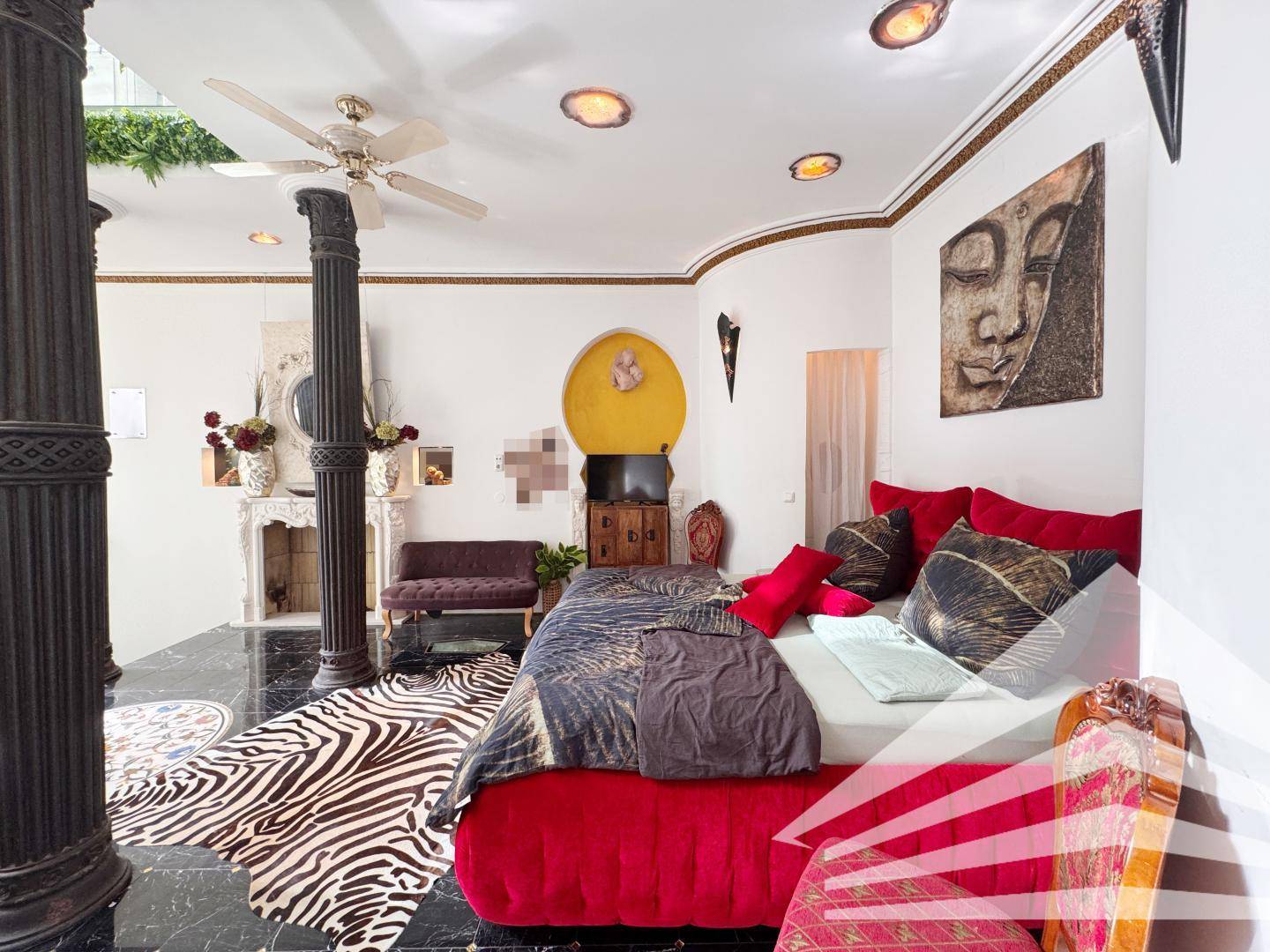 Exotisch gestaltetes Schlafzimmer mit rotem Bett, Deckenventilator und dekorativem Kamin.