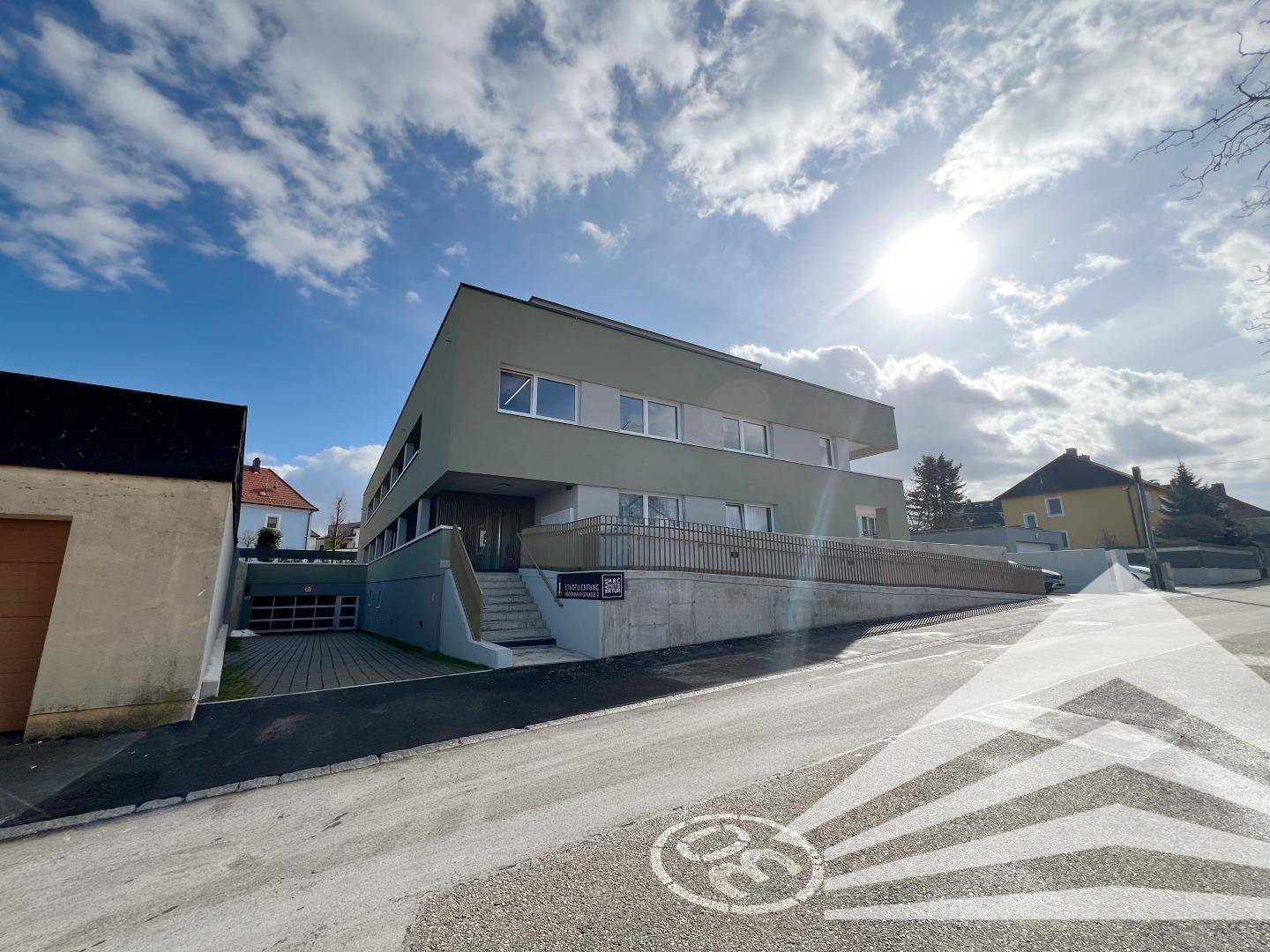 Moderne Wohnanlage mit heller Fassade, Eingangsbereich und Zufahrt von der Straße aus gesehen.