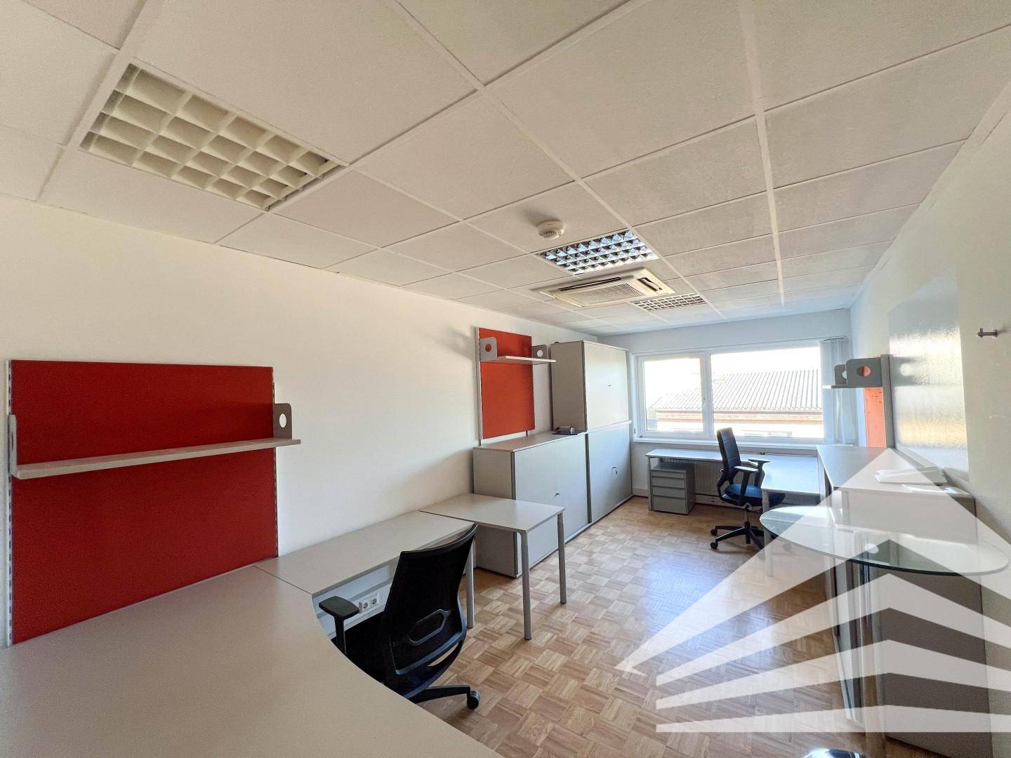 Büro mit mehreren Arbeitsplätzen, Schreibtischen, Bürostühlen, roten Akzenten und Fenstern.