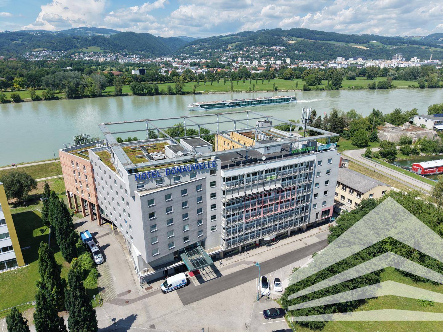Luftaufnahme des Hotels Donauwelle am Fluss mit grünen Dächern und umliegender Landschaft.