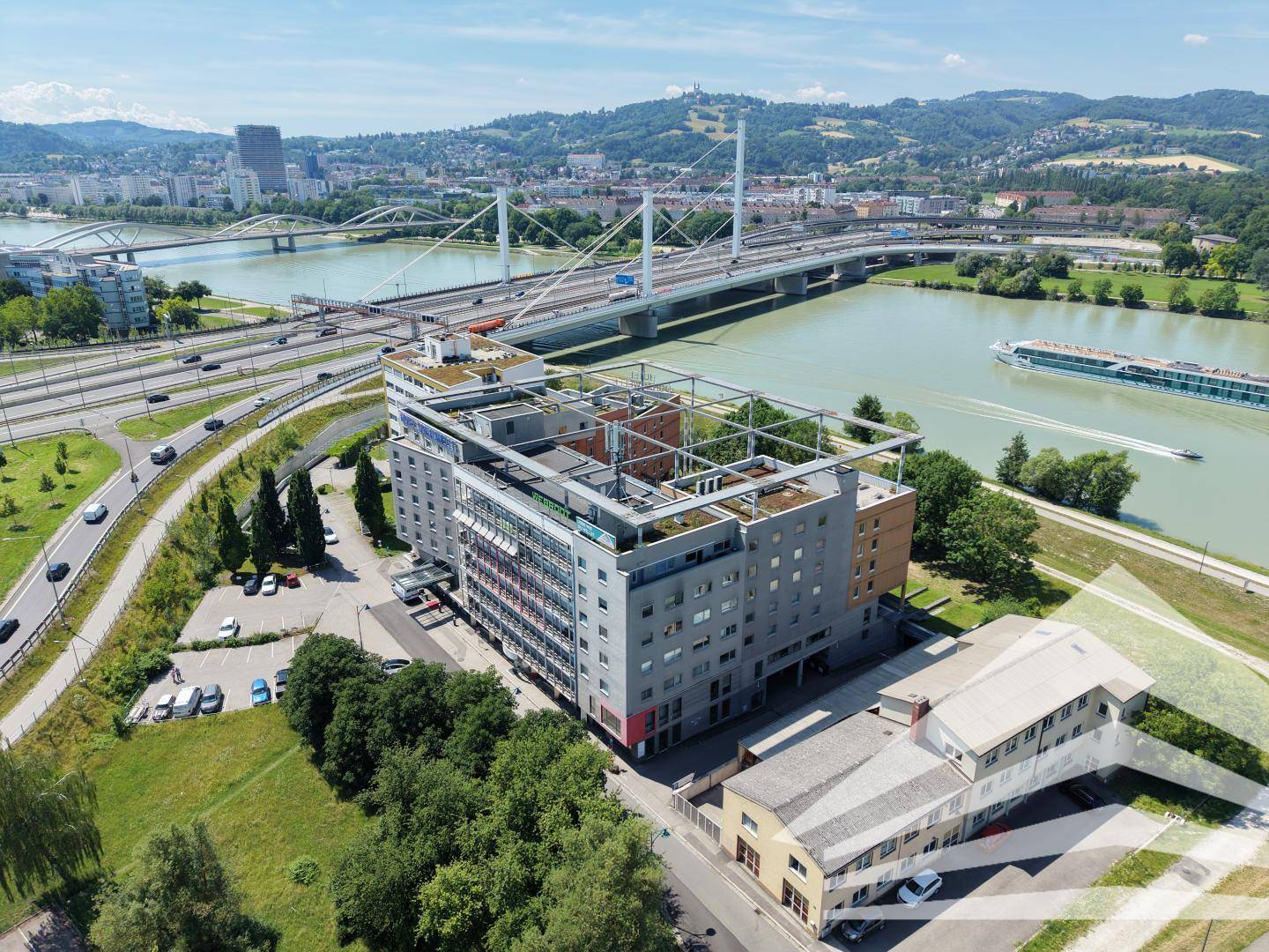 Luftaufnahme eines großen Hotelkomplexes am Flussufer mit Brücke und grüner Umgebung.